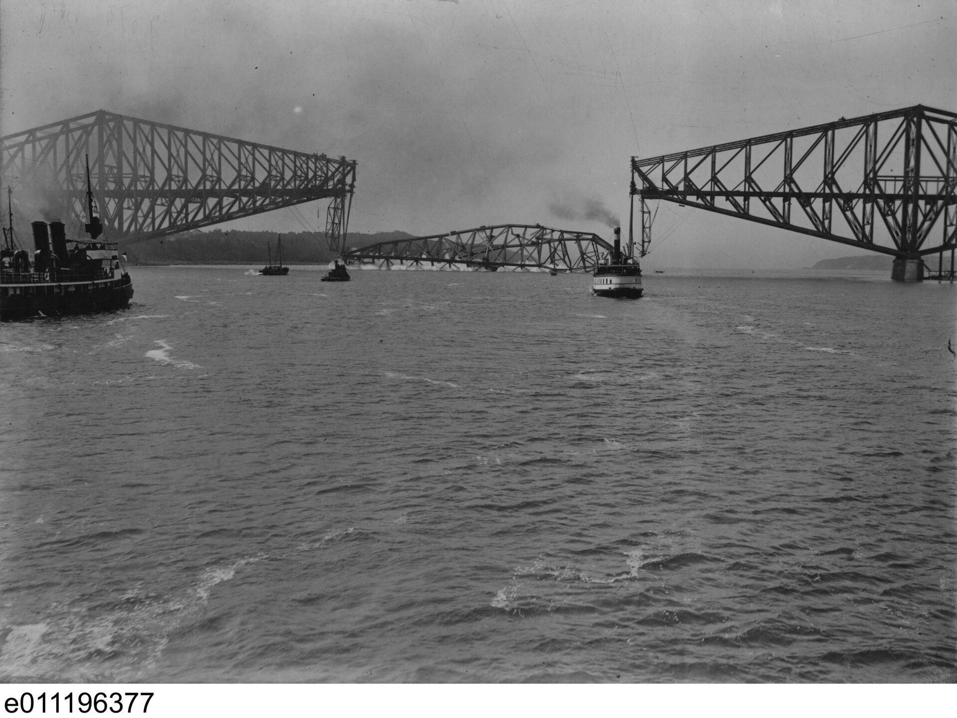The Quebec Bridge's central span lying in the St. Lawrence River between the two finished cantilever arms, with boats nearby.